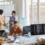 Time multidisciplinar de desenvolvimento trabalhando em escritório moderno com computadores e código na tela