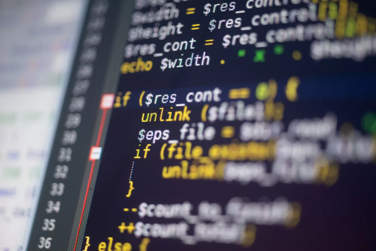 A macro close-up of a monitor showing colorful source code (PHP) on a dark background. The focus is sharp in the center, highlighting commands like "if" and "unlink," with the rest slightly blurred.