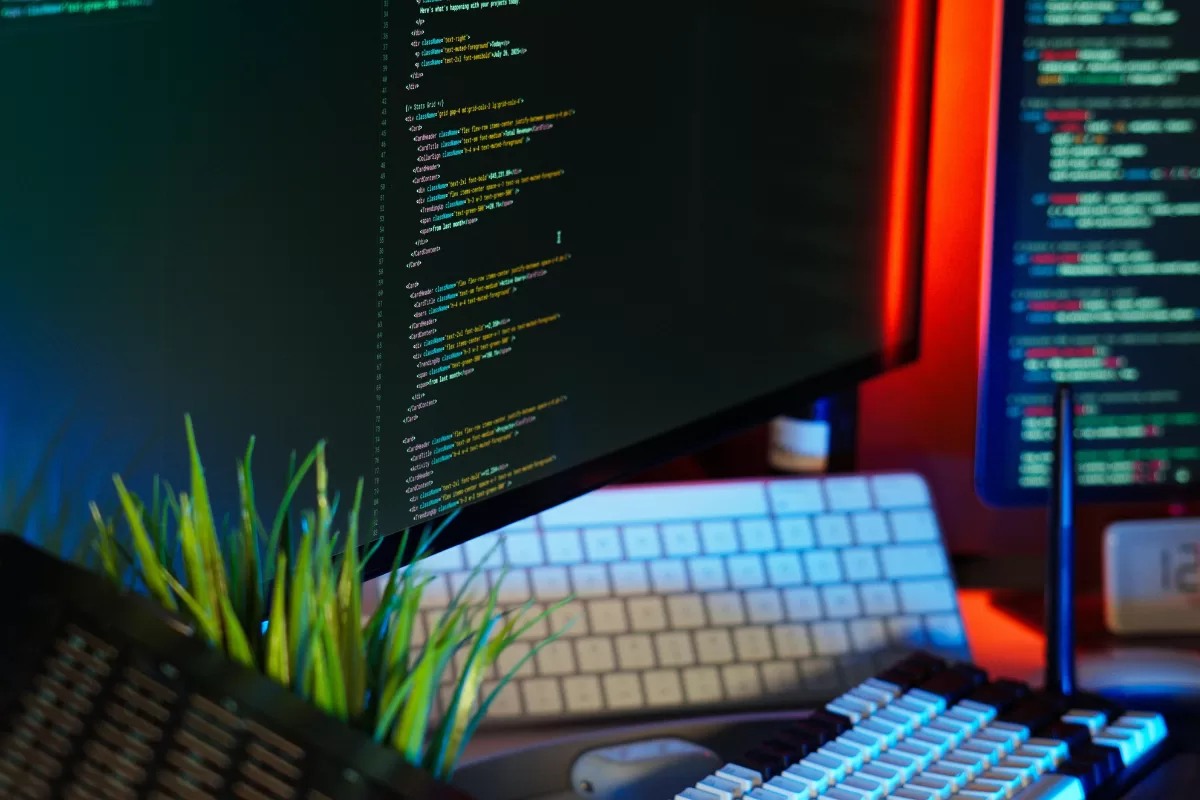 Monitor de computador exibindo código de programação, teclado, mouse e planta próximos, ambiente iluminado com luzes coloridas.