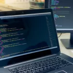 Um notebook aberto em uma mesa de trabalho exibindo código HTML/CSS em uma tela escura. Ao lado, há um segundo monitor também mostrando códigos, representando um ambiente de desenvolvimento de software profissional.