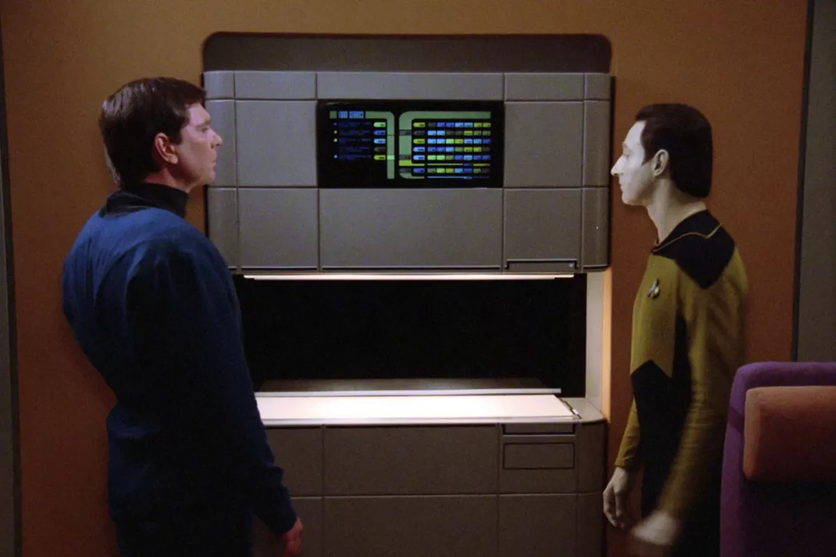 Dois personagens observam um replicador futurista em funcionamento na série Star Trek.