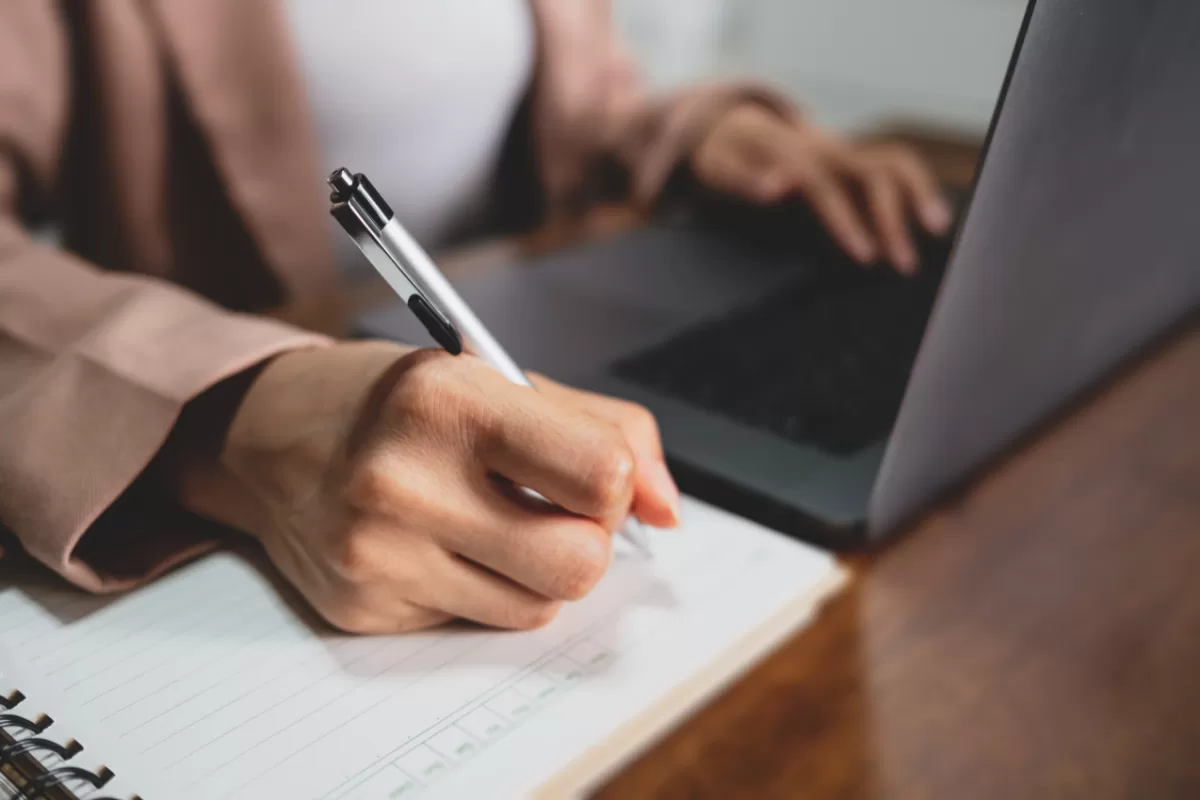 Person writing in spiral notebook with silver pen while working on laptop in professional work environment