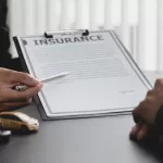 Profissional de seguros apresentando contrato de apólice em prancheta para cliente assinar, com miniatura de carro ao lado simbolizando seguro automotivo