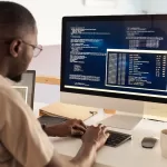 Engenheiro de software trabalhando com código em tela de computador, analisando dados e desenvolvendo sistemas em ambiente de trabalho moderno