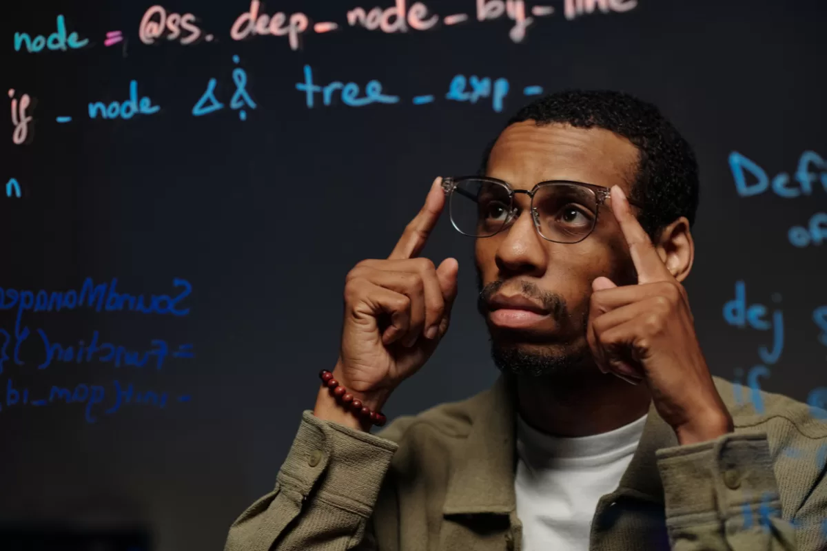 Um homem negro com óculos de armação fina olha para cima com uma expressão reflexiva e concentrada, tocando as têmporas com os dedos indicadores. Ao fundo, um painel de vidro escuro contém anotações técnicas e fragmentos de lógica de programação escritos à mão em cores vibrantes como azul, laranja e branco.