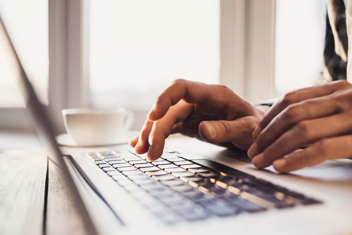 A close-up focus shot of a person's hands typing on a silver laptop keyboard. The setting is softly lit by natural light from a window in the background, where a blurred white coffee cup is visible.