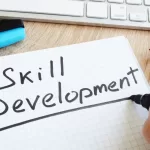Mão escrevendo "Skill Development" em caderno sobre mesa de trabalho com teclado, simbolizando aprendizado contínuo em tecnologia