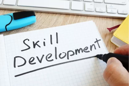 Mão escrevendo "Skill Development" em caderno sobre mesa de trabalho com teclado, simbolizando aprendizado contínuo em tecnologia