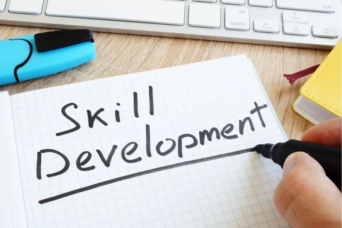 Mão escrevendo "Skill Development" em caderno sobre mesa de trabalho com teclado, simbolizando aprendizado contínuo em tecnologia