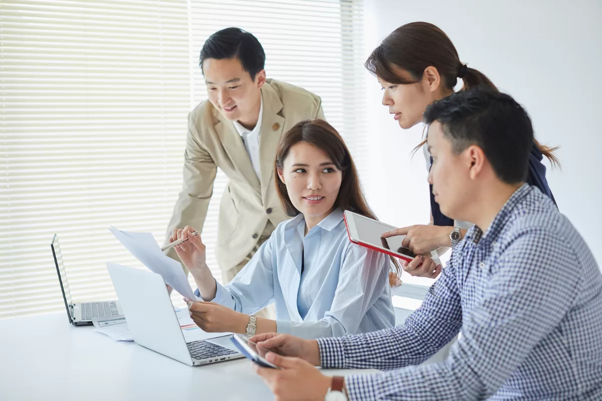 Four professionals in a collaborative work meeting around a white table. A woman points at a laptop screen while her colleagues observe, use a tablet, and analyze documents.