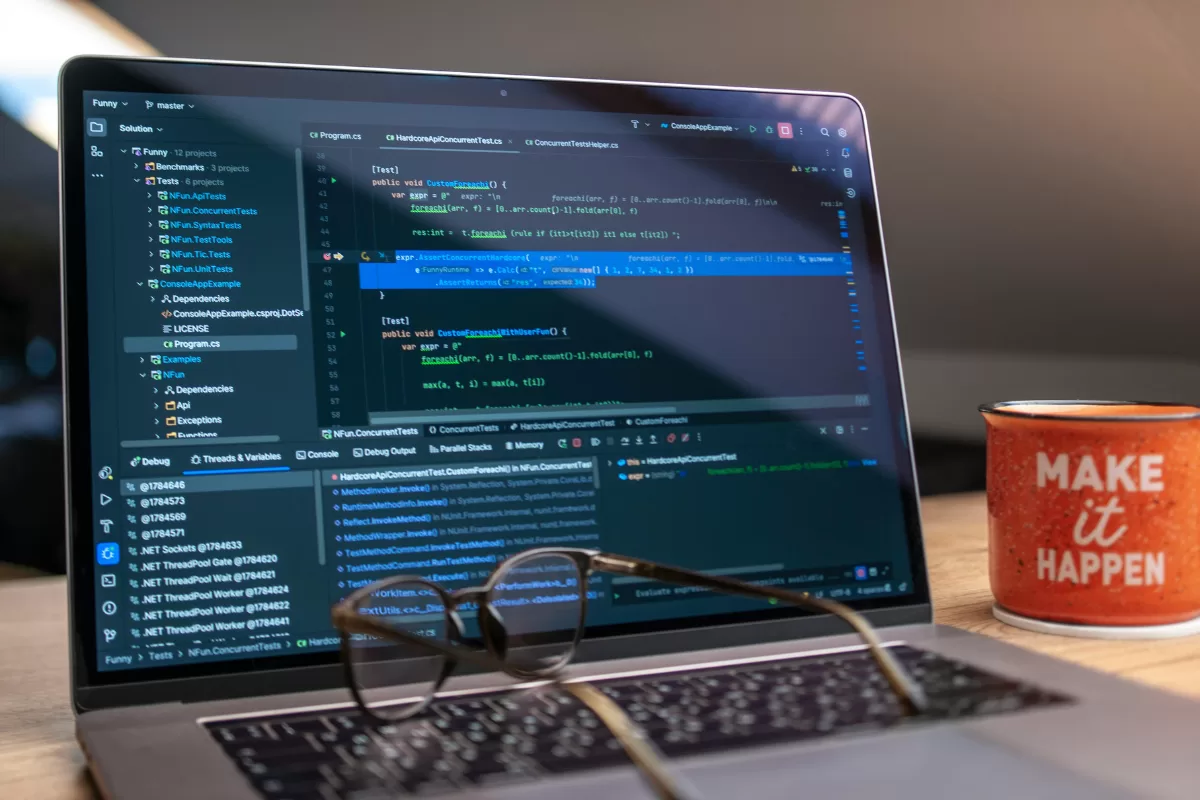 View of an open laptop screen displaying lines of programming code in an Integrated Development Environment (IDE). A pair of glasses sits on the keyboard, and an orange mug with the phrase "Make it happen" is next to it. 