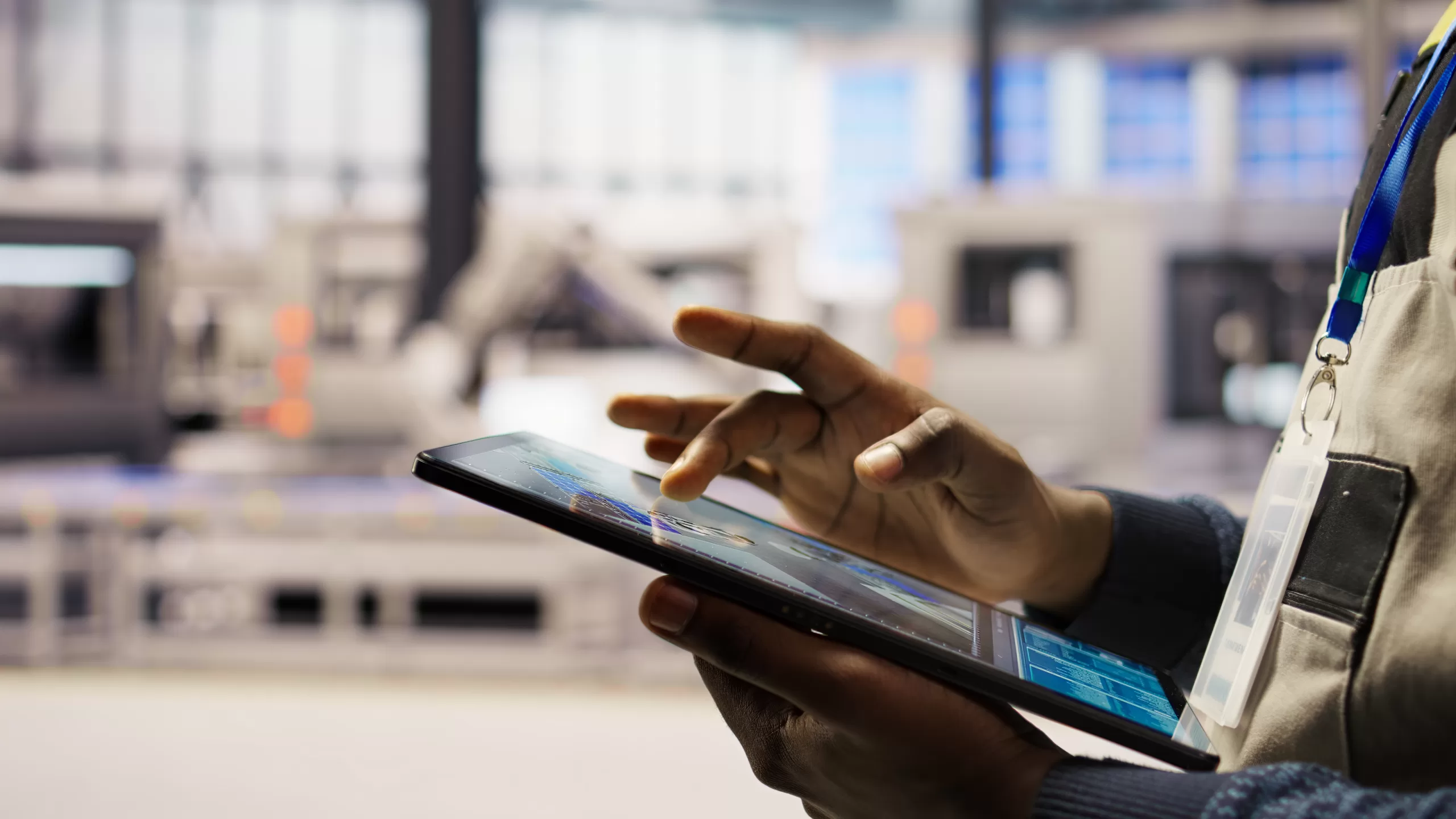 Close-up of an engineer's hands using a digital tablet inside a factory. The screen displays technical blueprints and layout data. The background is blurred, showing a clean, high-tech industrial environment.