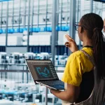 Dois engenheiros em uma fábrica inteligente. Em primeiro plano, uma mulher negra com tranças segura um notebook que exibe um modelo 3D de braços robóticos. Ao fundo, um colega observa a linha de produção automatizada repleta de maquinário moderno.