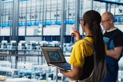 Dois engenheiros em uma fábrica inteligente. Em primeiro plano, uma mulher negra com tranças segura um notebook que exibe um modelo 3D de braços robóticos. Ao fundo, um colega observa a linha de produção automatizada repleta de maquinário moderno.