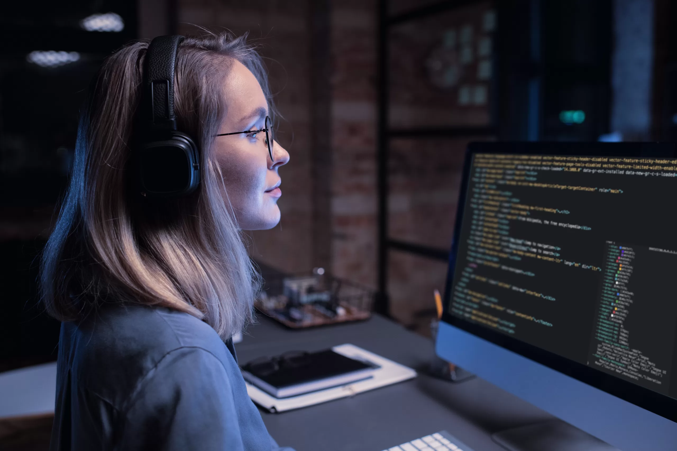 A young woman in profile wearing large headphones and glasses, looking focused at a computer monitor in a dark room. The screen displays colorful lines of programming code.