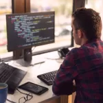 Desenvolvedor full stack trabalhando em código no monitor enquanto utiliza laptop, representando o dia a dia de um profissional de tecnologia