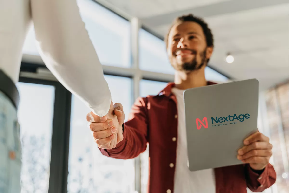 Smiling professional shaking hands with partner while holding tablet with NextAge logo, representing software development partnership