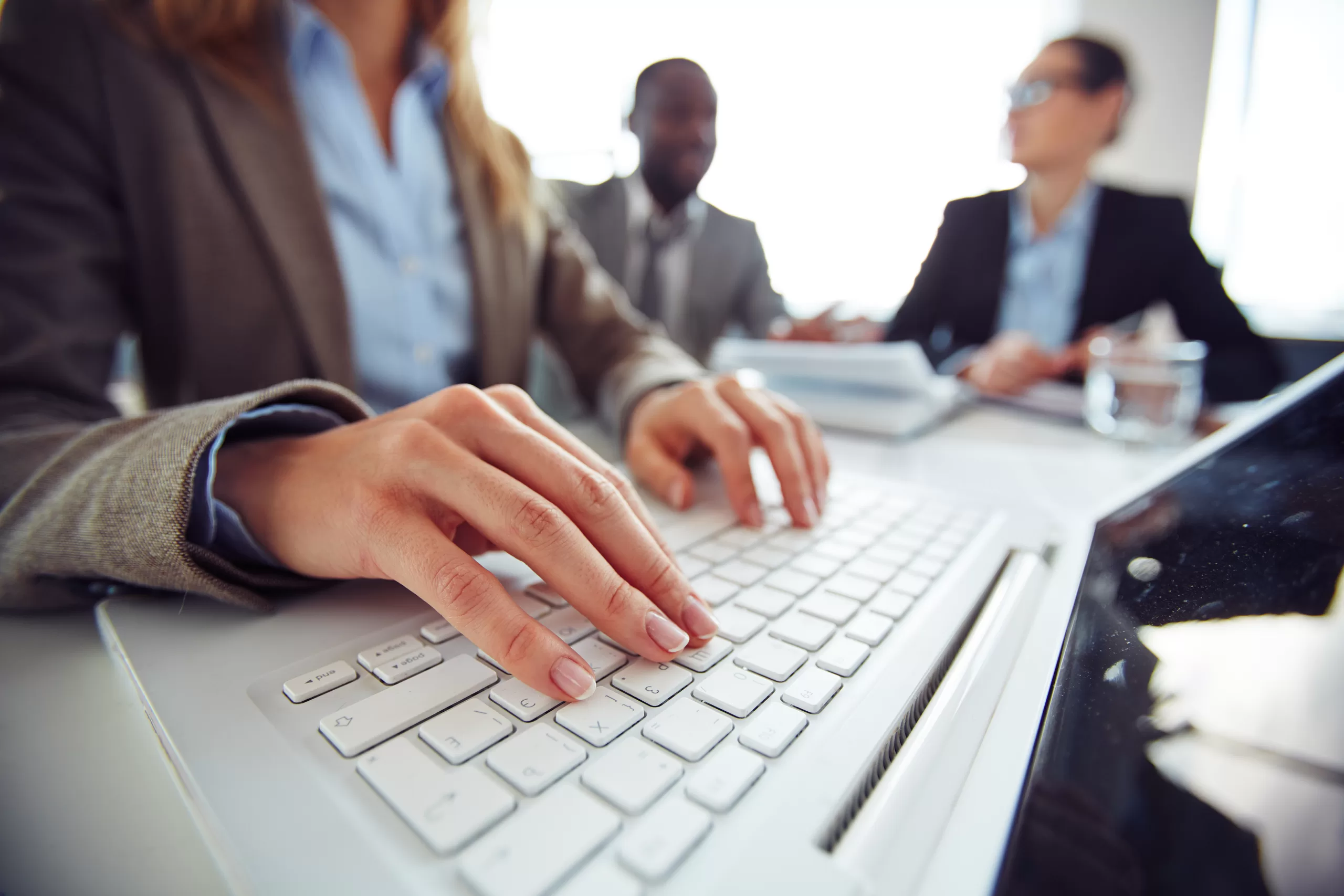 Professional typing on laptop during business meeting with team in the background in corporate environment