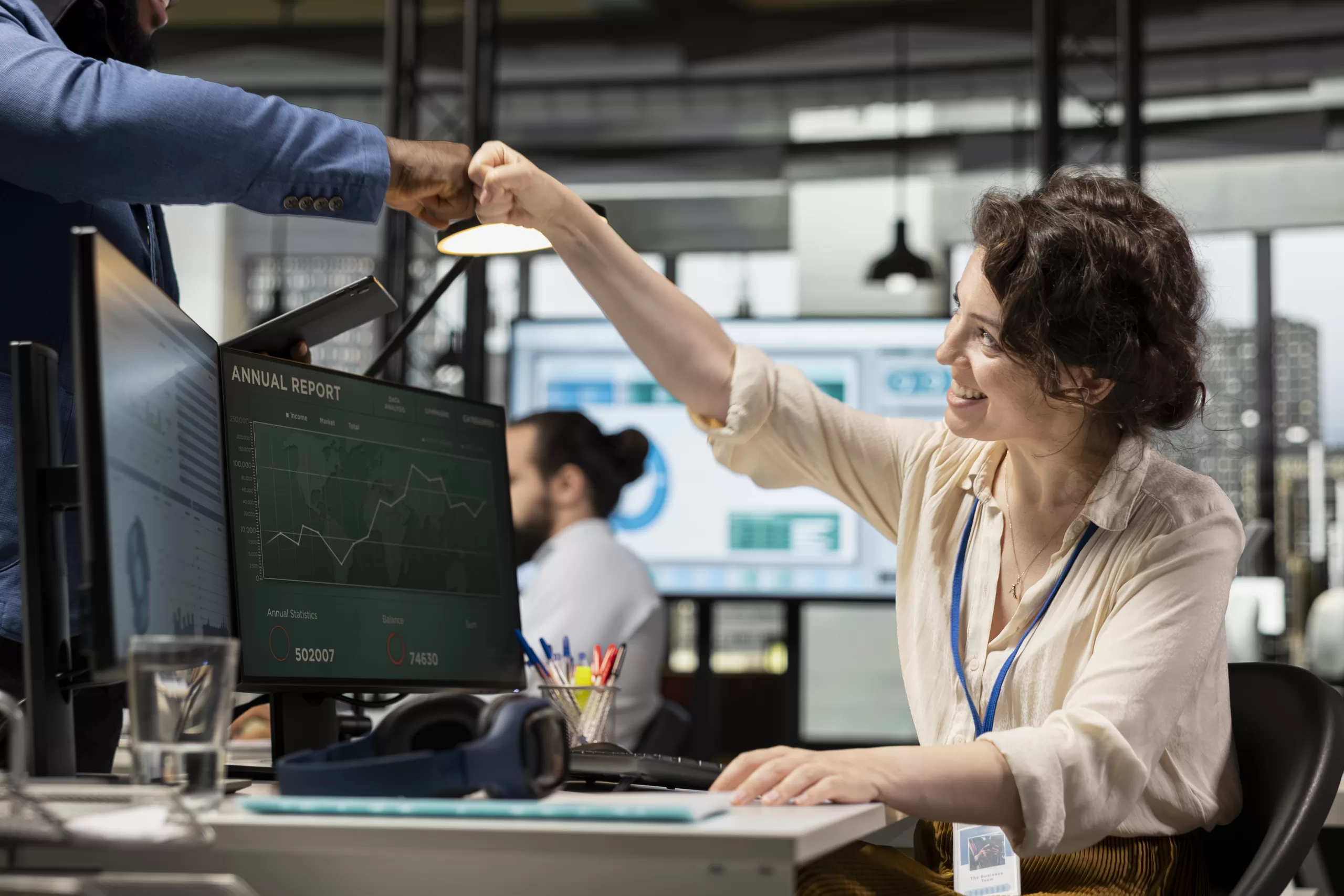 Coworkers greeting each other with fist bump in modern office with data and graphs on computer screen