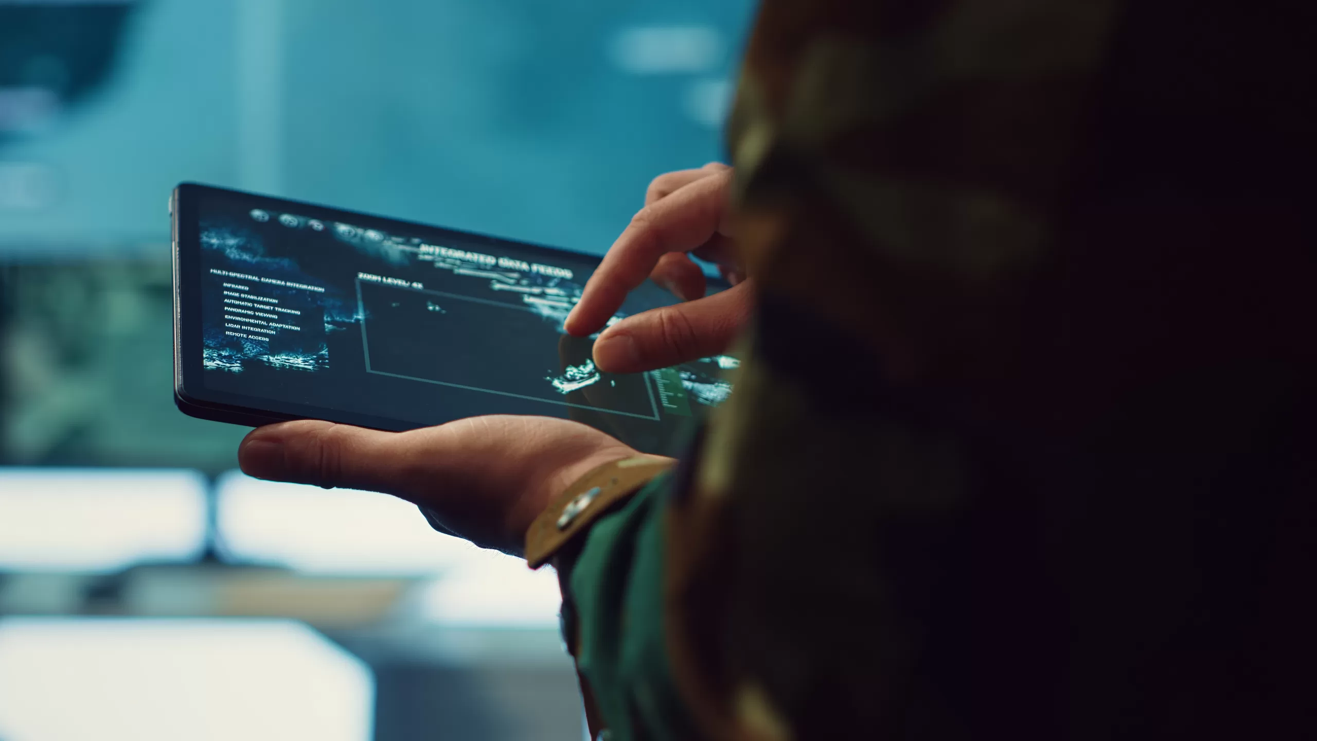 Person holding a tablet displaying an integrated data feed interface in a monitoring environment with screens in the background.