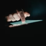 Mão tocando a tela iluminada de um tablet em ambiente escuro, representando o uso de aplicativos mobile