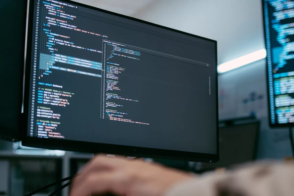 Monitor displaying lines of code in a dark text editor, with a person typing in the background in a multi-screen setup