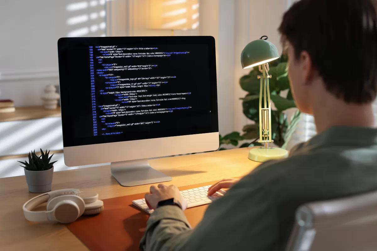 Developer typing code on an iMac monitor displaying HTML syntax highlighted in blue, in a warmly lit home office setup.