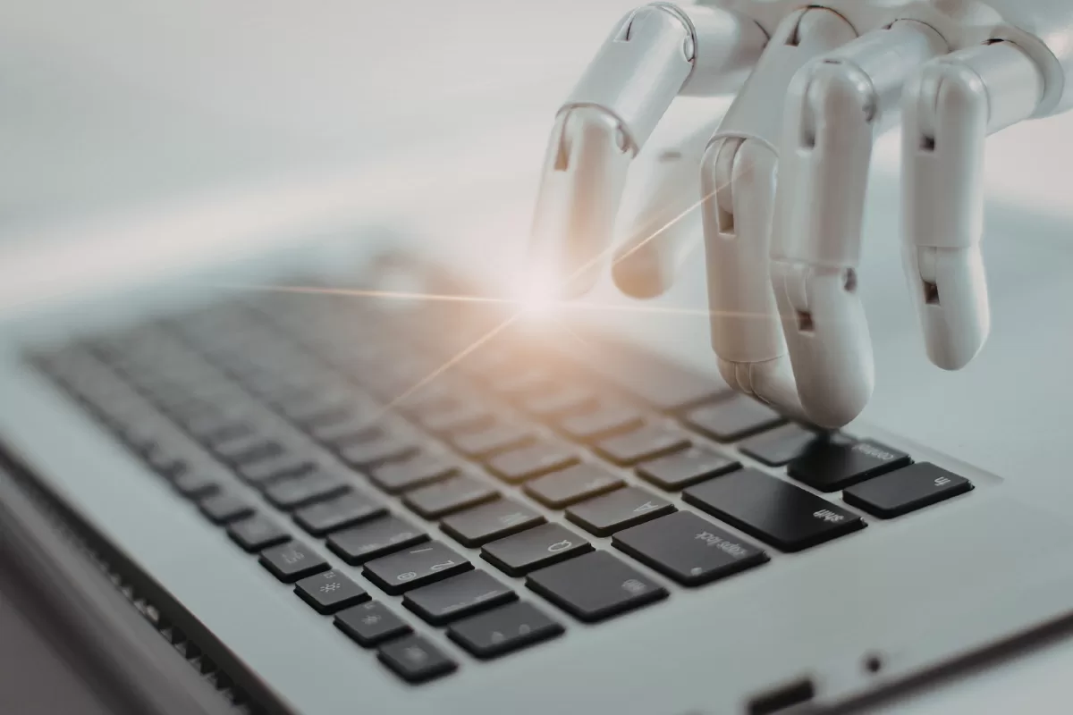 A detailed, white articulated robotic hand hovering over an open laptop keyboard. Its fingers are in a typing pose. A bright flare of light emanates from where the fingers are about to press the spacebar.