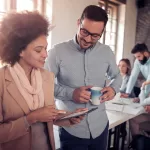 Dois profissionais em pé revisam informações em um tablet em um ambiente de escritório, enquanto outros colegas trabalham ao fundo.