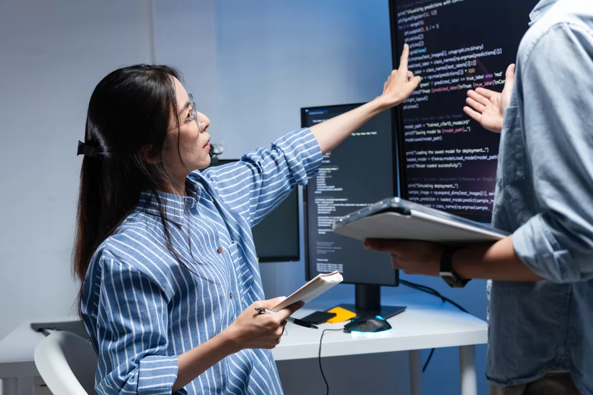 Uma desenvolvedora com óculos e camisa listrada aponta para uma tela vertical de monitor com código de programação, discutindo o projeto com um colega que segura um tablet. O ambiente é um escritório de tecnologia moderno com iluminação técnica azulada ao fundo.