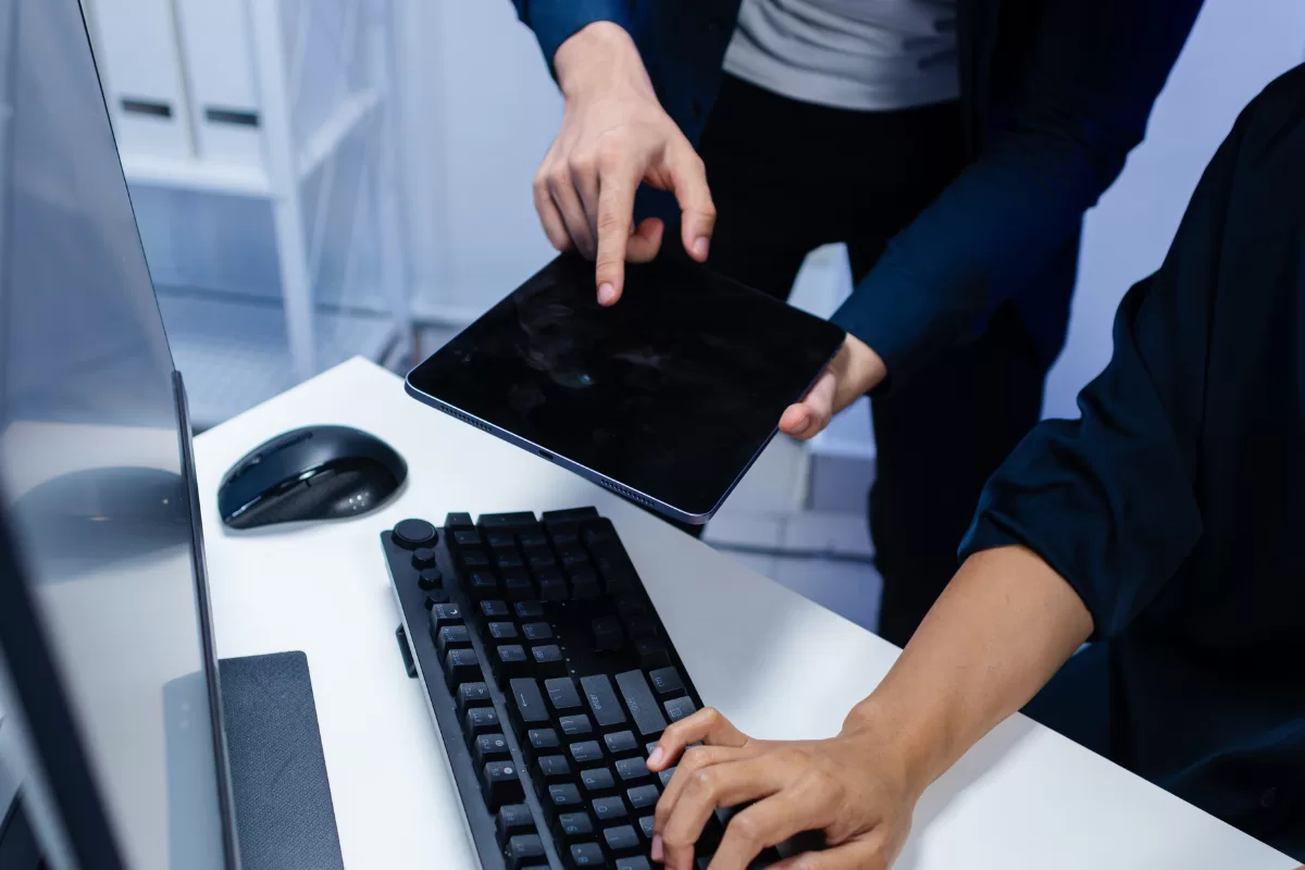 An overhead view of two people at a modern office workstation. One person is seated, hands resting on a black keyboard. A second standing person holds a tablet (screen is black) and points to it with a finger, sharing something. A PC monitor and mouse are on the white desk. The lighting is cool and blue.