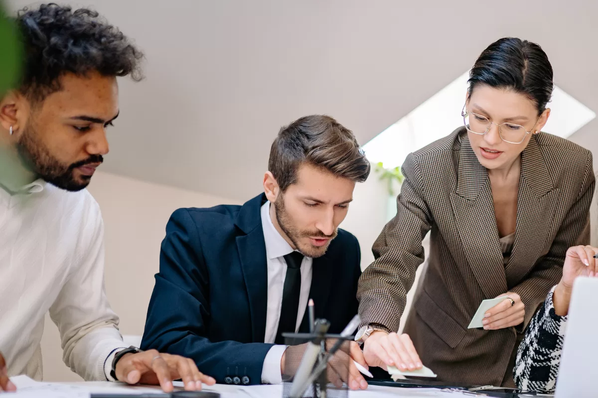 Team of professionals gathered around a table reviewing printed charts and reports, with open laptops and documents spread across the desk.