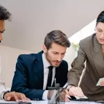 Equipe de profissionais reunida em torno de uma mesa analisando gráficos e relatórios impressos, com laptops abertos e documentos espalhados sobre a mesa.