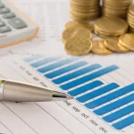 Relatório financeiro com gráfico de barras azul, caneta e moedas empilhadas ao lado de uma calculadora, simbolizando análise e gestão financeira