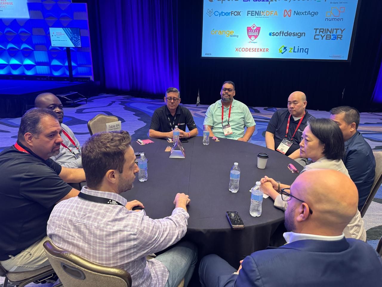 Attendees engaged in a roundtable discussion at the Midsize Enterprise Summit Spring 2026, with the NextAge logo displayed among the event's sponsor brands in the background.