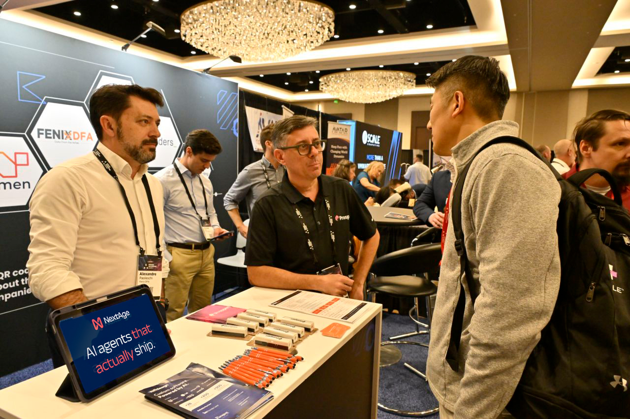 NextAge representative talking with attendees at the company's booth at the Midsize Enterprise Summit Spring 2026, with a tablet displaying the message "AI agents that actually ship