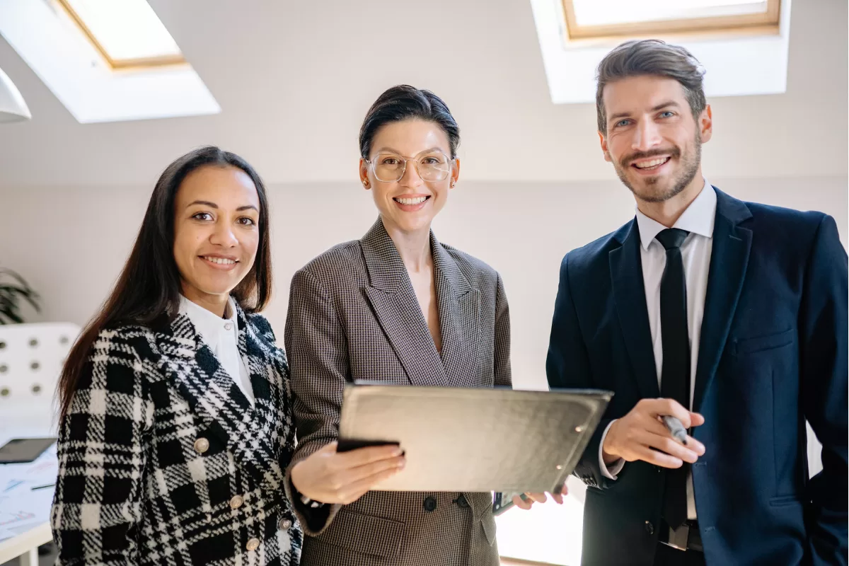 Three business professionals smiling in an office setting, holding a tablet, representing an aligned and collaborative outsourcing team.