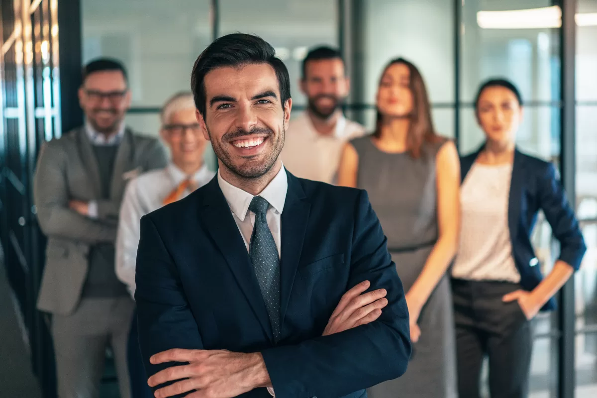 Team leader smiling with arms crossed, with a group of professionals in the background, representing a dedicated development squad led by an experienced Tech Lead.