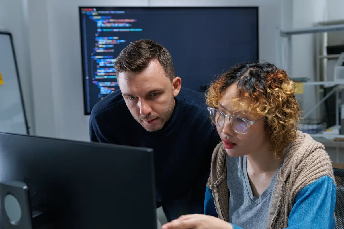 Two developers reviewing code on a monitor, with a colorful code screen in the background, illustrating the technical work of a software development squad.