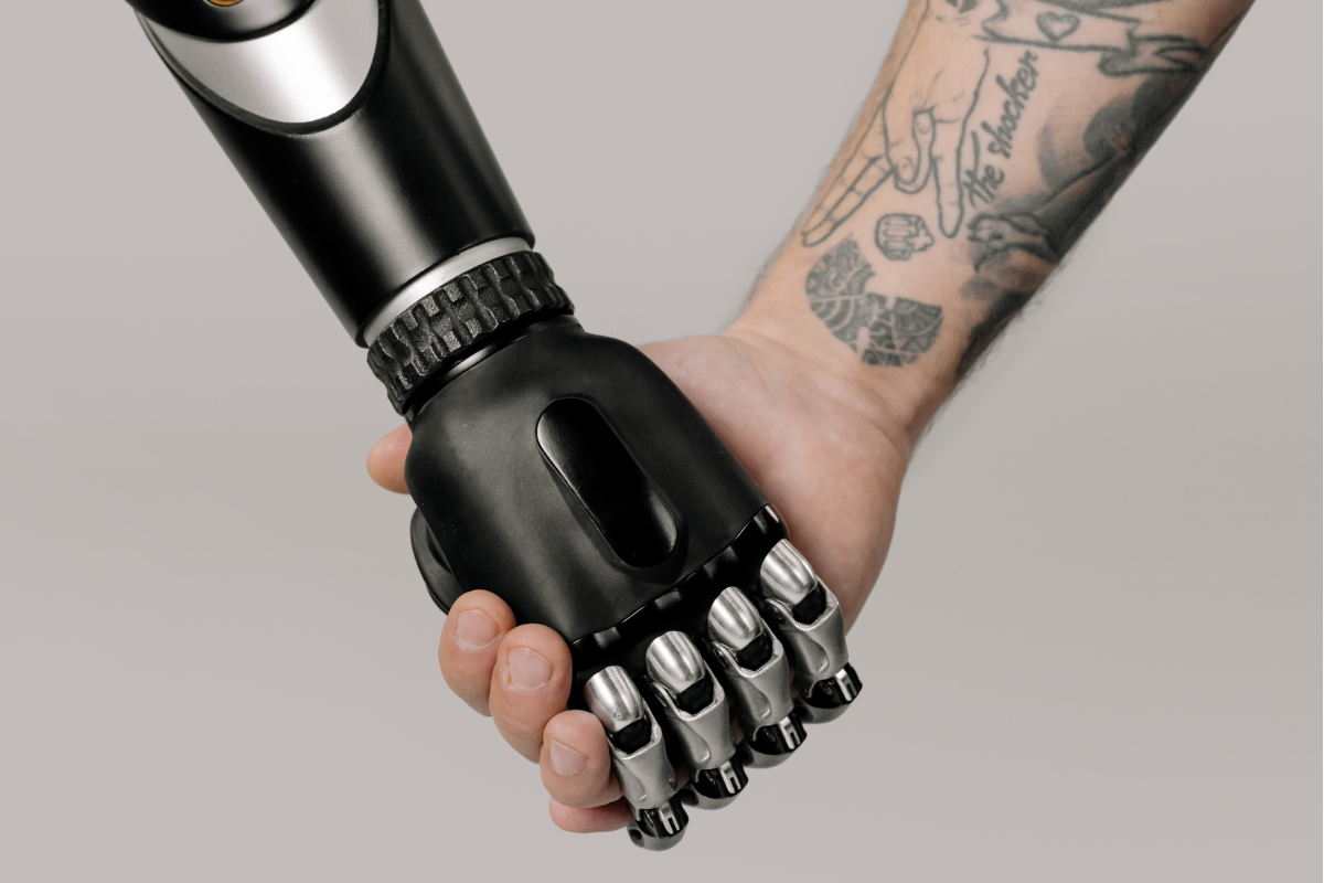 Mão humana com tatuagens segurando uma mão robótica preta e prateada, simbolizando a colaboração entre humanos e máquinas