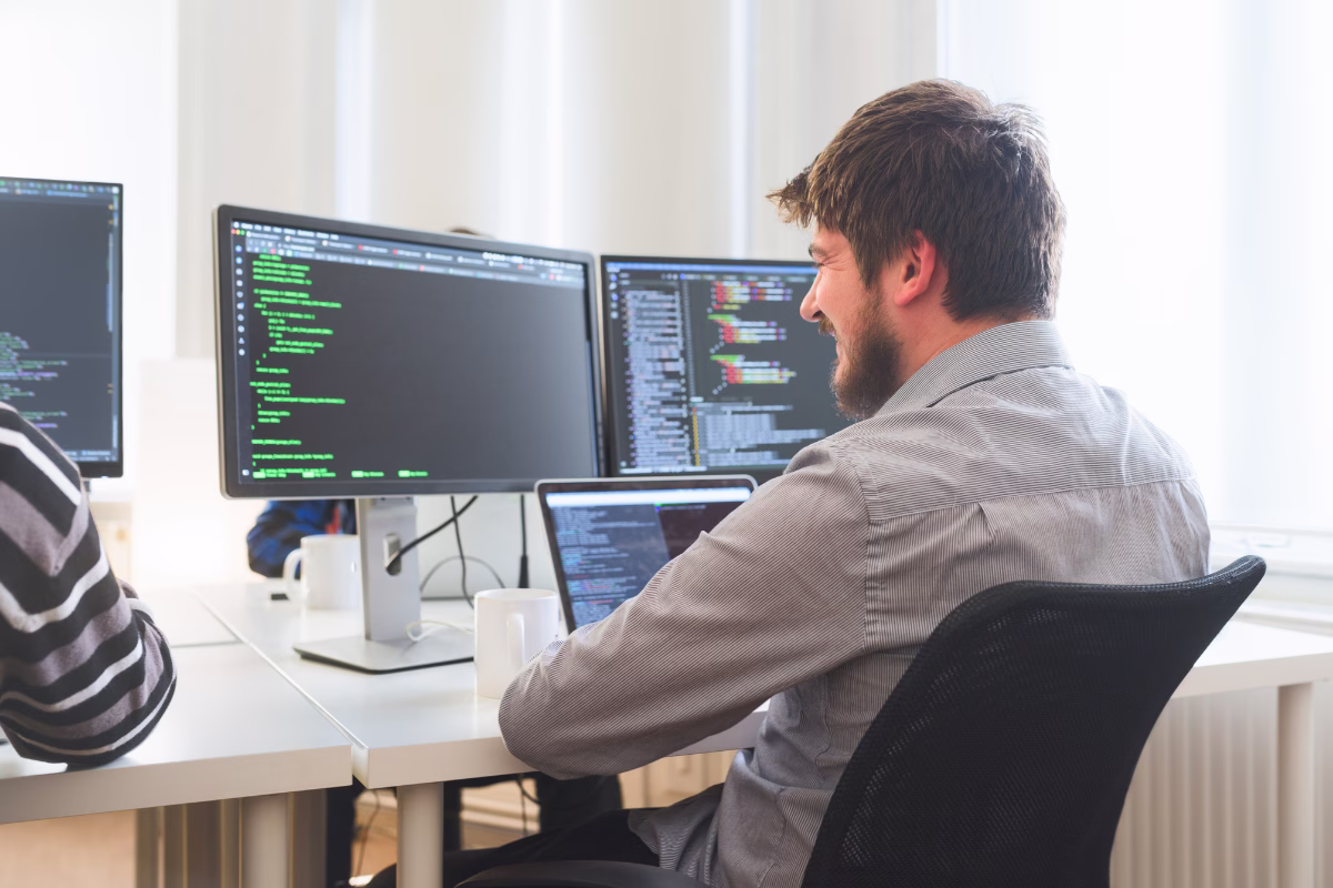 Desenvolvedor de software trabalhando com múltiplos monitores exibindo código, ilustrando a escassez de profissionais de TI nas empresas