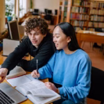 Uma foto colorida mostra dois estudantes jovens, um rapaz e uma moça, sentados lado a lado em uma mesa de madeira dentro de uma biblioteca bem iluminada.