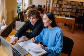 Uma foto colorida mostra dois estudantes jovens, um rapaz e uma moça, sentados lado a lado em uma mesa de madeira dentro de uma biblioteca bem iluminada.