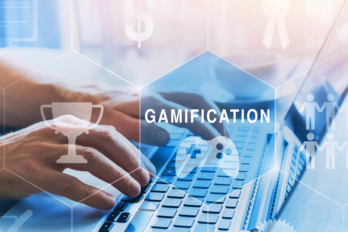 Close-up of a person's hands typing on a laptop keyboard. Overlaying the image is a translucent blue graphical design with hexagonal shapes containing icons of a trophy, a video game controller, and people. In the central hexagon, the word "GAMIFICATION" is prominently displayed in white capital letters.