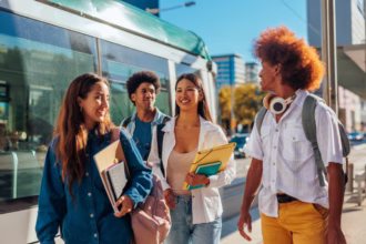 A imagem mostra um grupo diversificado de quatro jovens estudantes caminhando juntos ao ar livre em uma rua urbana ensolarada, ao lado de um veículo de transporte público