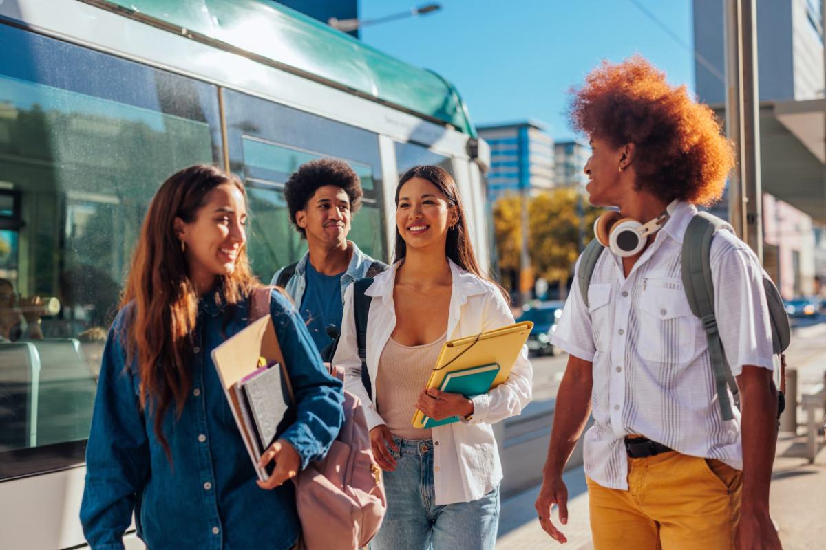A imagem mostra um grupo diversificado de quatro jovens estudantes caminhando juntos ao ar livre em uma rua urbana ensolarada, ao lado de um veículo de transporte público