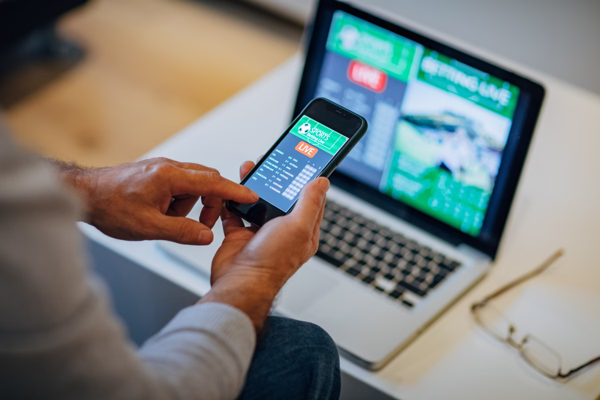 A close-up shot, focused on a person's hands holding a smartphone, with an index finger pointing to the phone's screen. The smartphone displays a live sports betting scoreboard and table with the text "SPORTS Betting LIVE" at the top.