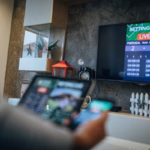 Uma imagem de primeiro plano, desfocada, mostra a parte de trás da cabeça e do ombro de uma pessoa enquanto ela olha para uma televisão grande de tela plana. Na tela da TV, uma interface de apostas ao vivo está visível com um placar de 2-1 entre Portugal e Austrália, e o texto "BETTING LIVE" em cima.