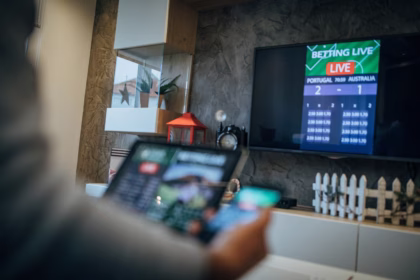 Uma imagem de primeiro plano, desfocada, mostra a parte de trás da cabeça e do ombro de uma pessoa enquanto ela olha para uma televisão grande de tela plana. Na tela da TV, uma interface de apostas ao vivo está visível com um placar de 2-1 entre Portugal e Austrália, e o texto "BETTING LIVE" em cima.