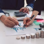 Dois profissionais analisando gráficos financeiros em uma mesa com calculadora, moedas empilhadas e notas, representando a análise de custo de desenvolvimento de software